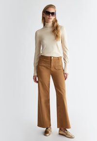 Maglione aderente beige a collo alto abbinato a pantaloni marroni a gamba larga con vita alta, dotati di tasche testurizzate. Il modello indossa occhiali da sole scuri e scarpe dorate.