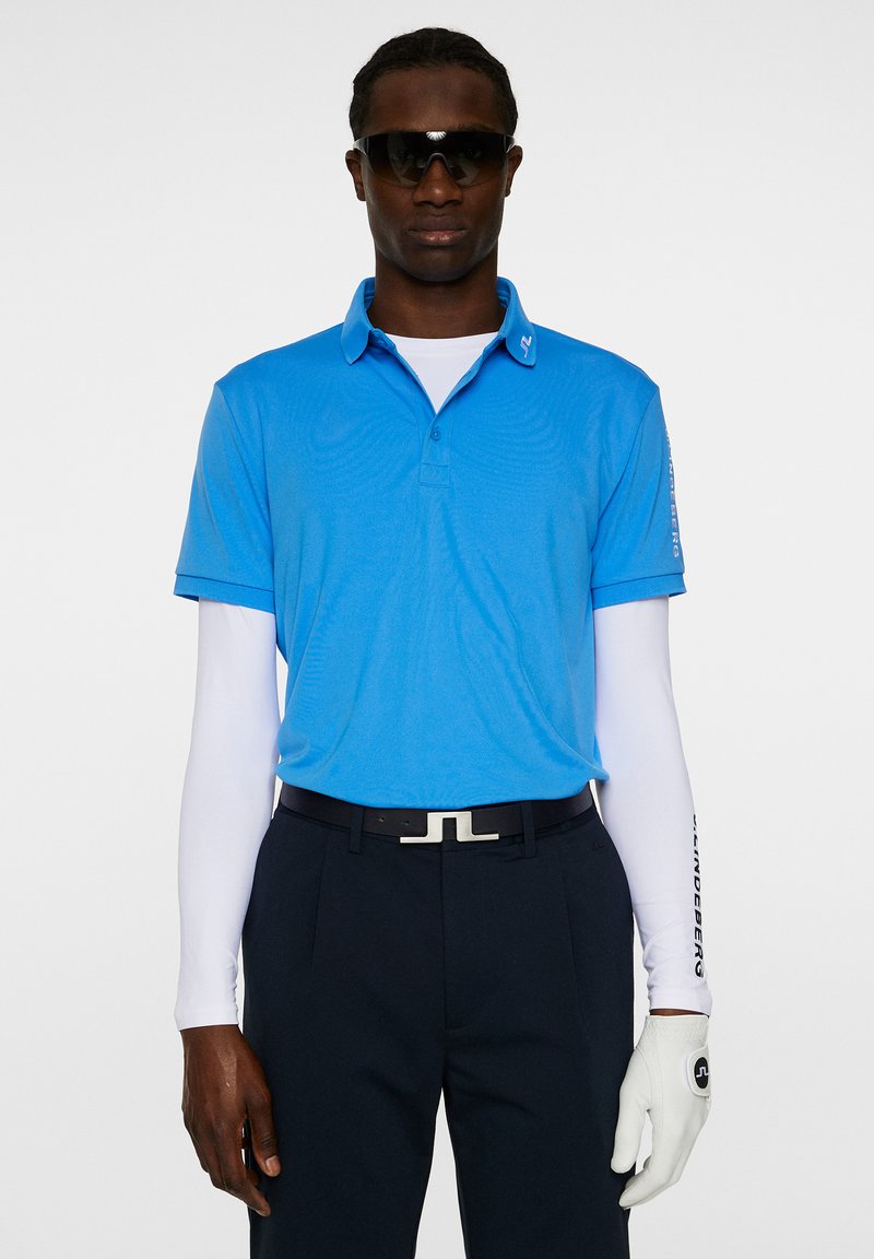 Sininen polo-paita, jossa on kaulus, yhdistettynä valkoiseen pitkään hihaiseen paitaan, musta vyö, tummat housut ja valkoinen käsine. Mustat aurinkolasit.