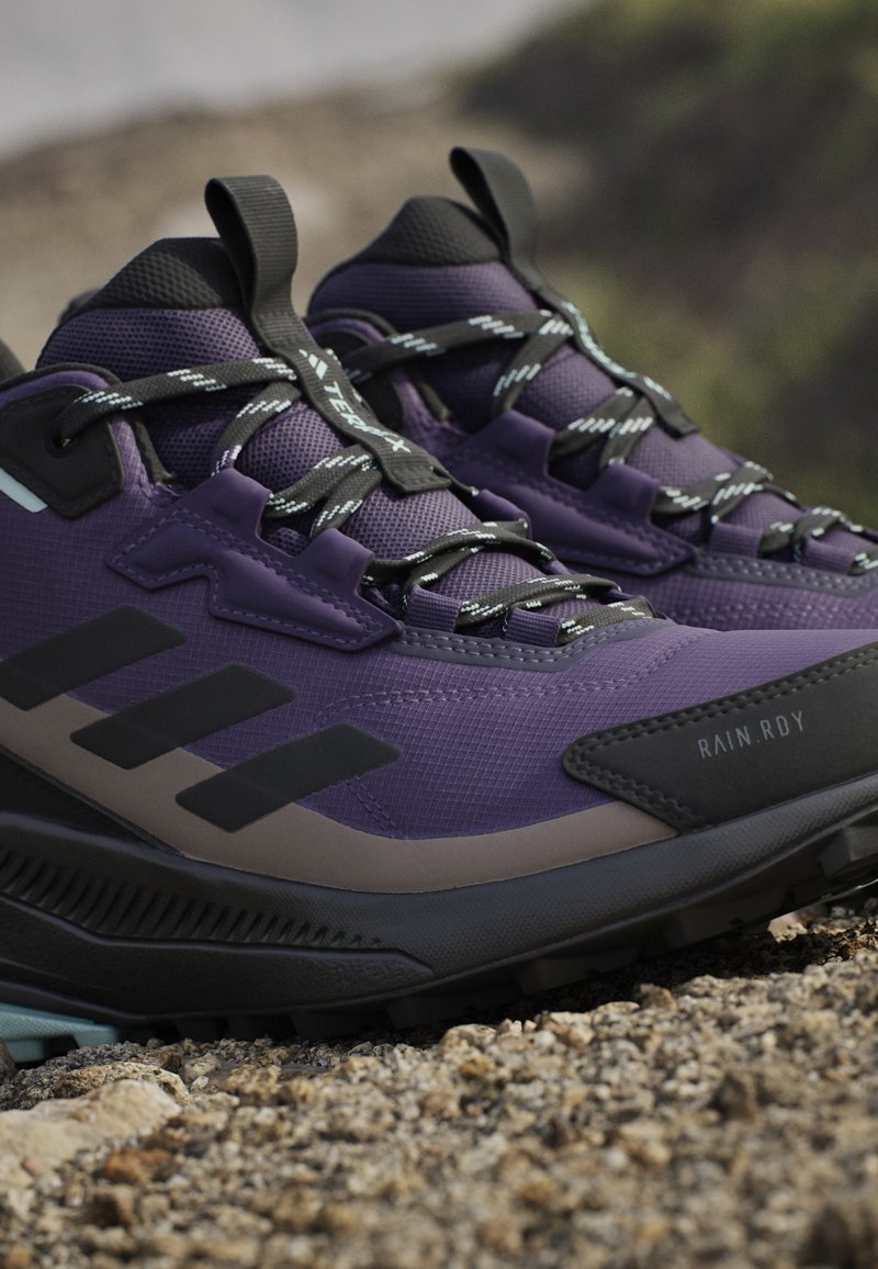 Zapatos de senderismo impermeables morados con acentos negros, superficie texturizada y cordones. Presentan una suela gruesa y marcas de la marca prominentes.