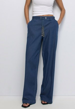 Femme portant un pantalon en jean bleu à jambes larges avec une taille à cordon et un débardeur blanc court, debout les mains dans les poches.