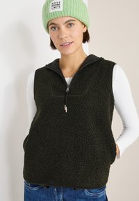 Fleeceweste in Dunkelgrün mit schwarzem Reißverschlusskragen, ausgestattet mit Fronttaschen und einer strukturierten, weichen Oberfläche. Trägt man über einem weißen Langarmshirt.