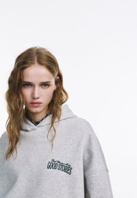 Felpa oversize grigia con cappuccio, caratterizzata da un testo ricamato che dice "Good Stories" in verde scuro. Tessuto morbido con un design casual.