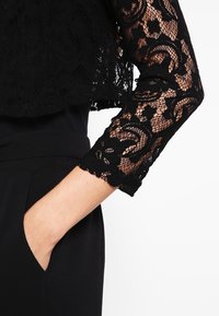 Svart spetskort topp med långa ärmar, med blommönster och scallopkanter. Bärs över ett enfärgat svart plagg, vilket visar en hand i fickan.