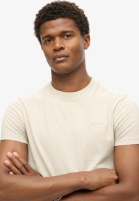 Beige bomulds t-shirt med rund halsudskæring, kortærmede og et lille broderet logo på brystet; glat tekstur, normal pasform.