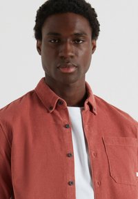 Camicia di velluto corduroy rossa con colletto button-down, tasca frontale e bottoni a contrasto, indossata sopra una maglietta bianca.