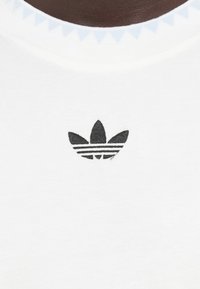 Λευκό βαμβακερό T-shirt με μαύρο λογότυπο Adidas με τρεις ρίγες, τοποθετημένο στο πάνω μέρος του στήθους, περιβαλλόμενο από μπλε κολάρο με υφή.