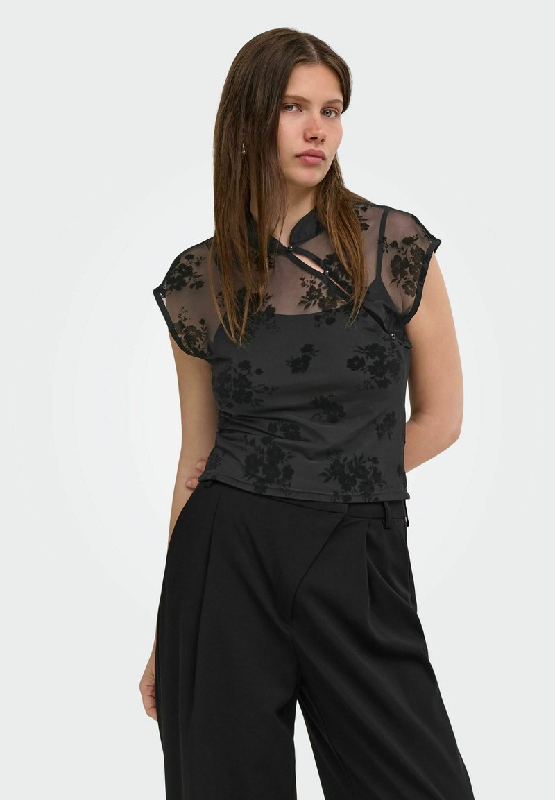 Jeune femme portant un top noir transparent à motifs floraux avec des manches courtes et un pantalon large noir taille haute, sur un fond clair uni.