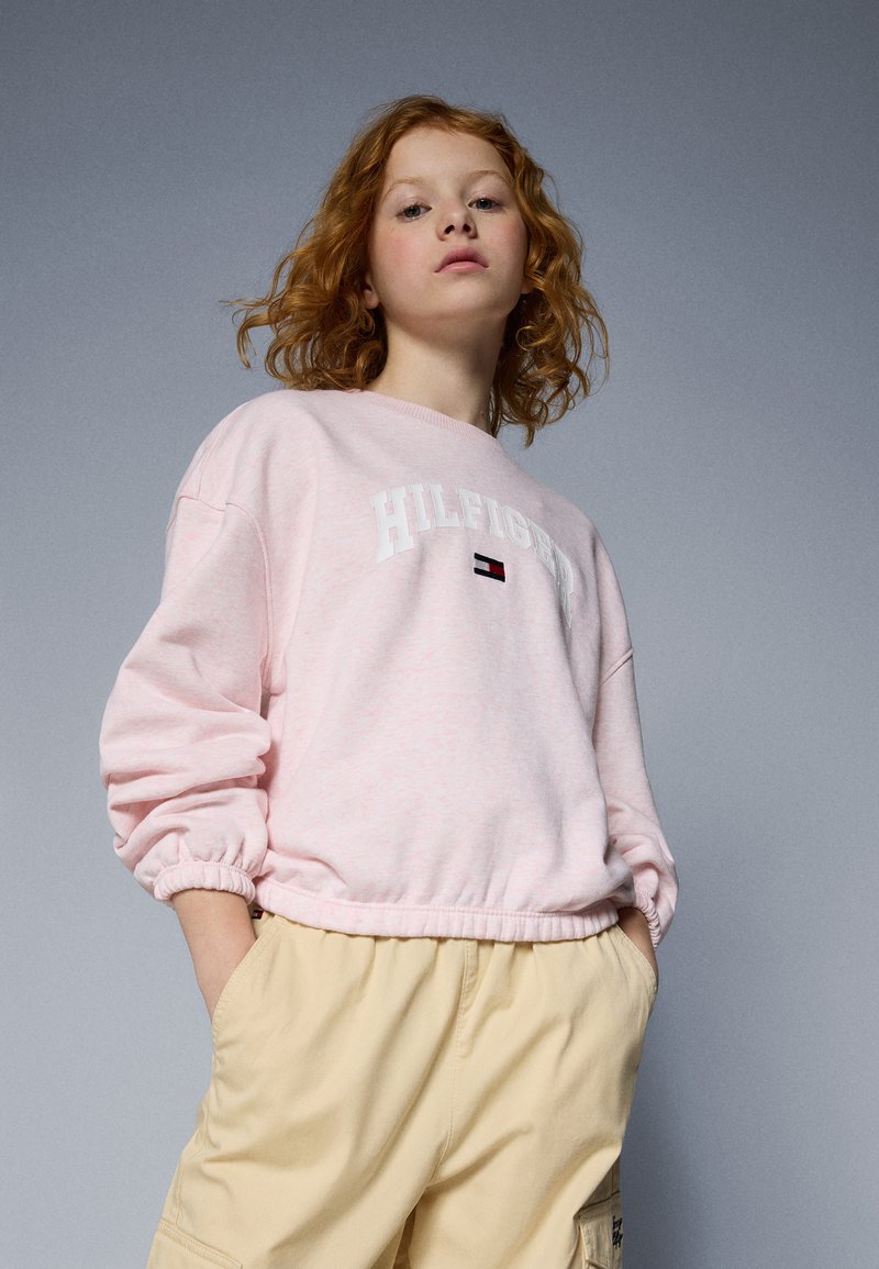 Sweatshirt rose clair avec des poignets et un ourlet côtelés, portant le texte blanc "HILFIGER" et un détail de logo rouge/blanc/bleu. Associé avec un short beige.
