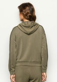 Donna con capelli castano mossi, indossa una felpa verde oliva e pantaloni coordinati, vista da dietro su uno sfondo bianco semplice.