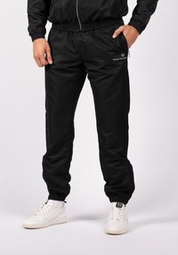 Svarta träningsbyxor i jogger-stil med elastisk midja, dragkedjefickor och en slät yta. Har en logotyp på vänster höft och muddar vid anklarna.