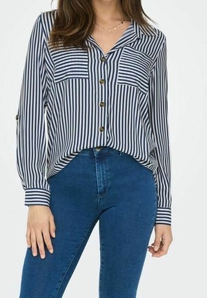 Vrouw draagt een marineblauw-wit verticaal gestreept overhemd met knopen, ingestopt in een hoog getailleerde blauwe spijkerbroek, met lange mouwen en borstzakken.