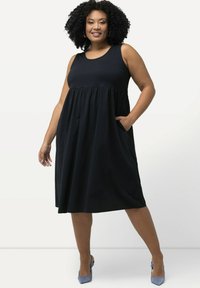 Vestito nero senza maniche con scollatura rotonda, vita arricciata, tasche laterali e tessuto morbido; modella che indossa tacchi azzurri.