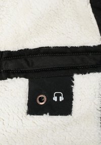 Svart stoff med lodden hvit fleece bakgrunn, med hodetelefonkontakt og et trykt hodetelefonikon. Kobberdetalj med øye er synlig.