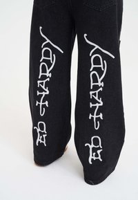 Jeans de mezclilla negra de pierna ancha con gran texto blanco "ED HARDY" impreso a lo largo de cada pierna. La tela tiene una textura suave y un diseño casual.
