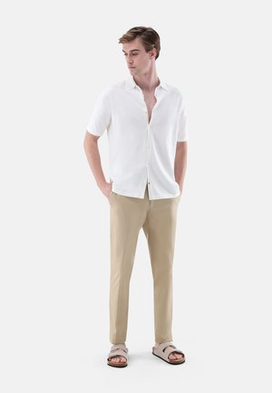 Junger Mann trägt ein weißes, kurzärmeliges Hemd mit Knöpfen, beige Hose und beige Sandalen, steht mit den Händen in den Taschen.