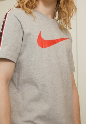 T-shirt en coton gris avec un logo Nike swoosh rouge bien en vue et des rayures noires sur les côtés avec des mini logos. Manches courtes, coupe décontractée.