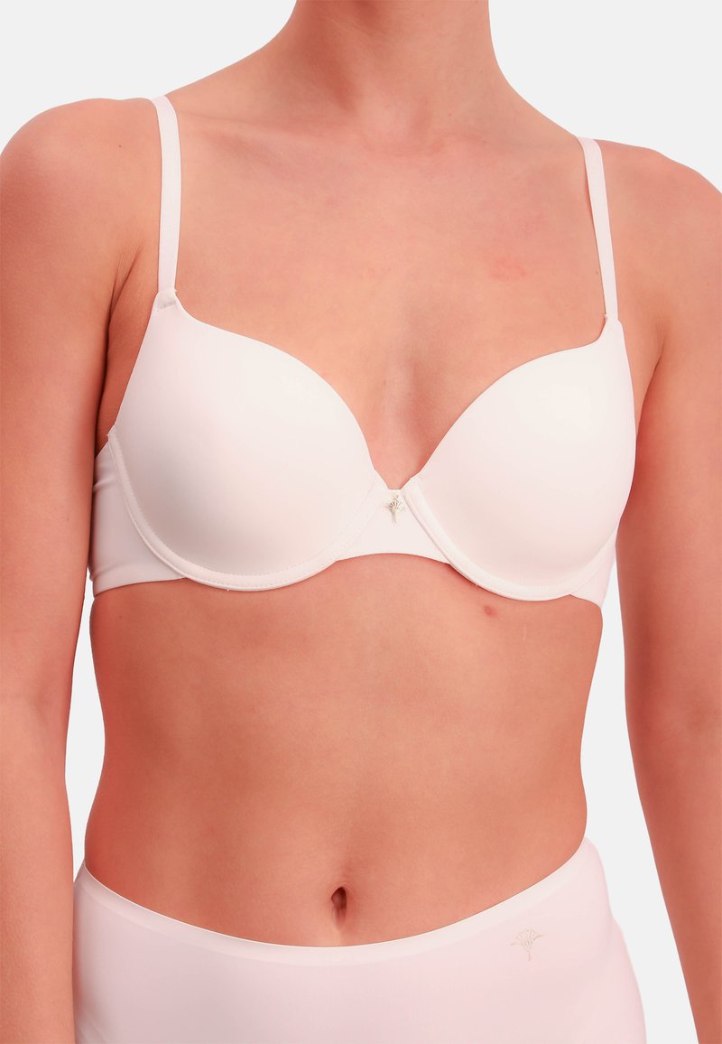 Femme portant un soutien-gorge blanc sans couture avec armatures et une culotte taille haute assortie sur un fond uni.