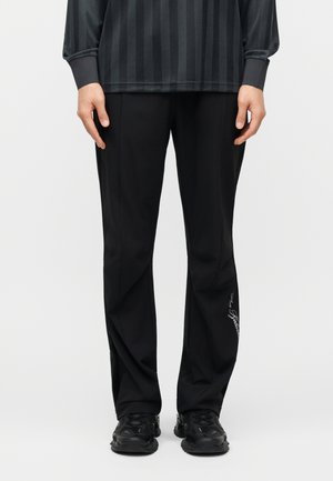 Personne portant un pantalon noir à jambes droites avec une petite inscription blanche sur la jambe gauche, associé à des baskets noires et un chemisier à manches longues rayé foncé.