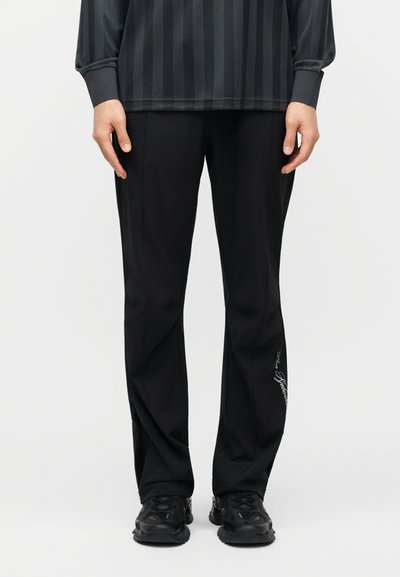 Personne portant un pantalon noir à jambes droites avec une petite inscription blanche sur la jambe gauche, associé à des baskets noires et un chemisier à manches longues rayé foncé.