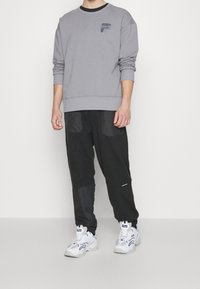 Šedá mikina s žebrovanými manžetami a lemováním, spárovaná s černými kalhotami s texturovanými panely a kapsami. Na sobě má bílé sportovní boty.