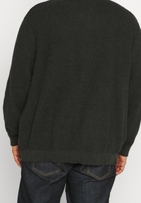 Dunkelgrüner strukturierter Strickpullover mit geripptem Saum, langen Ärmeln und lockerer Passform, kombiniert mit dunkelblauen Jeans mit Gesäßtaschen.