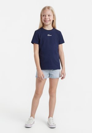 Niña sonriente de pie con una camiseta azul marino de Ellesse, pantalones cortos vaqueros claros y zapatillas blancas sobre un fondo blanco.