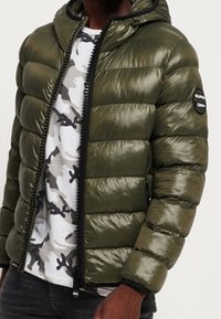 Chaqueta acolchada color verde oliva con textura brillante, capucha, cierre de cremallera y detalles en negro. Se lleva sobre una camisa con patrón de camuflaje gris.