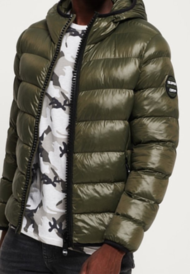 Chaqueta acolchada color verde oliva con textura brillante, capucha, cierre de cremallera y detalles en negro. Se lleva sobre una camisa con patrón de camuflaje gris.