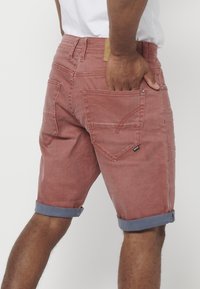 Shorts en denim rouge avec des poignets en bleu roulés, des poches avant et arrière, et une étiquette en cuir beige. Tissu texturé avec des détails de couture.