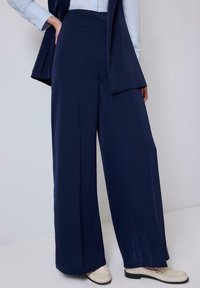 Wide-leg navy trousers made of soft material with a high waist. Features a smooth texture and clean lines, paired with light-colored shoes.