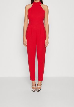 Femme portant une combinaison rouge sans manches à col halter avec un pantalon plissé et des escarpins argentés à bout pointu avec des brides à la cheville.