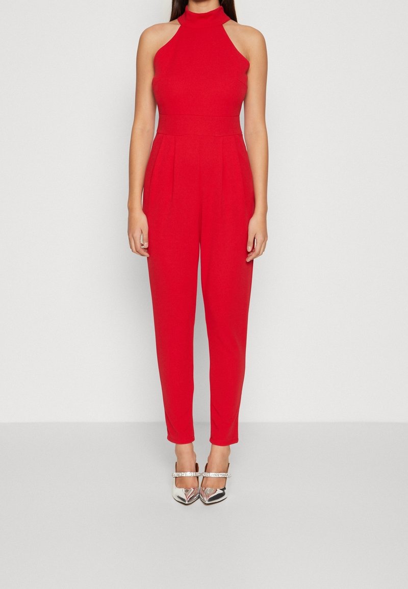 Femme portant une combinaison rouge sans manches à col halter avec un pantalon plissé et des escarpins argentés à bout pointu avec des brides à la cheville.