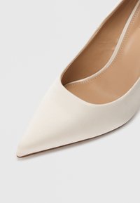 Γόβα με μυτερή μύτη από απαλό λευκό σατέν με λεία υφή και διακριτική λάμψη. Το εσωτερικό διαθέτει ελαφριά μπεζ επένδυση.