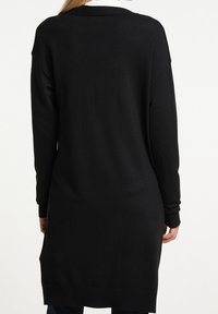 Osoba nosící dlouhý černý pletený svetr se dlouhými rukávy, zobrazena zezadu na jednobarevném bílém pozadí.