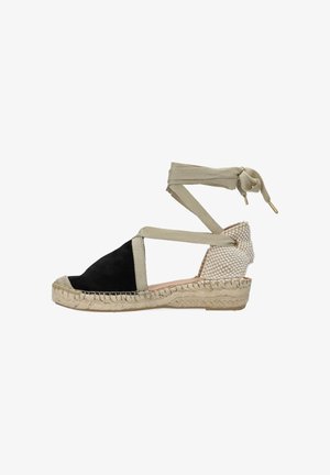 Cuña de espadrille de punta cerrada con parte delantera negra, talón tejido beige y lazos beige para atar al tobillo sobre fondo blanco.