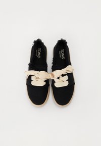 Scarpe slip-on in tela nera con bordi sfilacciati, caratterizzate da un grande fiocco beige per la chiusura e una suola in corda di juta, etichettate "TOMS."