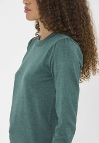 Teal tröja med rund halsringning, tillverkad av ett mjukt ribbat material. Har mjukt puffade ärmar och en slät yta med en figursydd design.