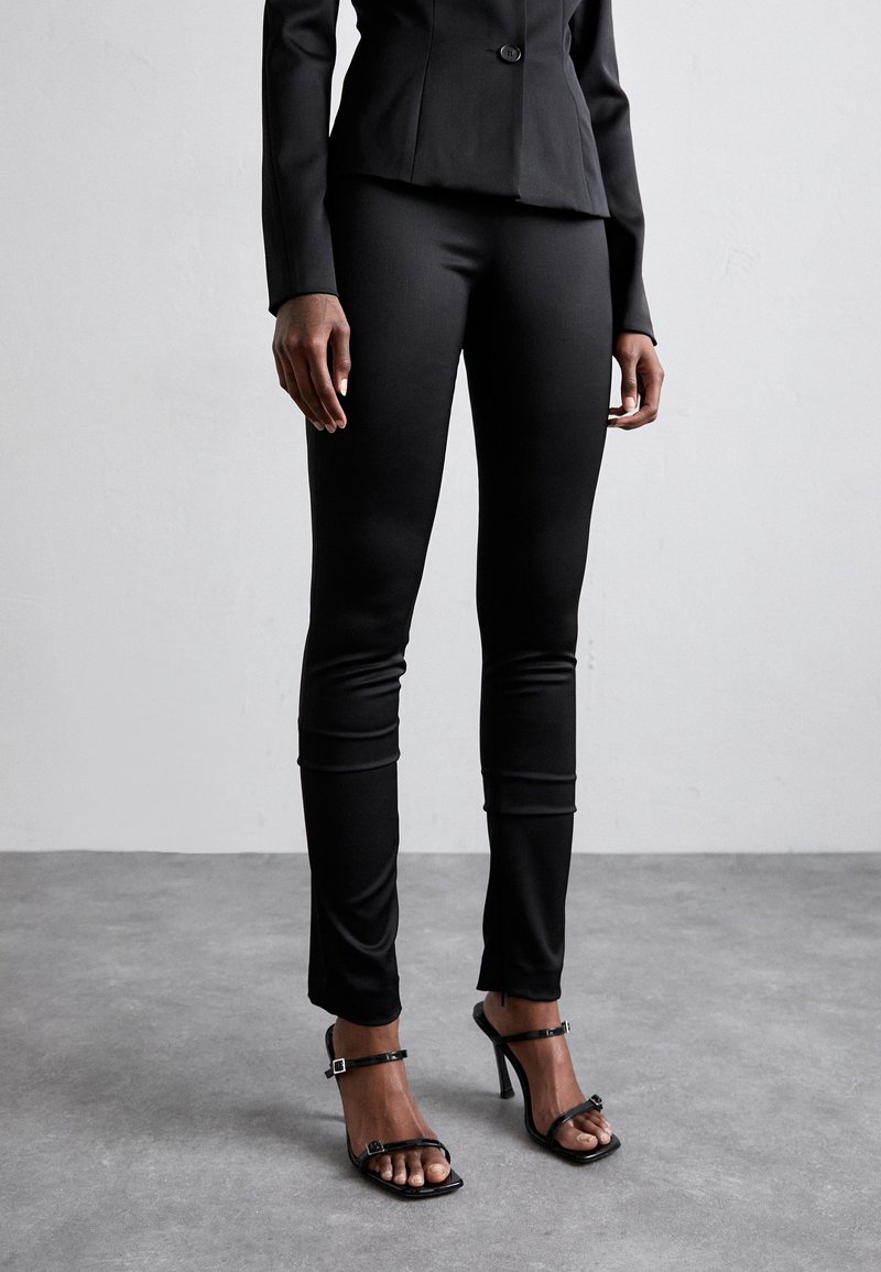 Personne portant un pantalon noir ajusté, un blazer noir et des sandales noires à talons hauts, debout sur un sol gris face à un mur clair et uni.