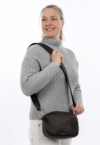 Braune Leder-Crossbody-Tasche mit genarbter Textur und Reißverschluss. Verstellbarer Riemen und silberne Beschläge. Getragen zu einem grauen Pullover.