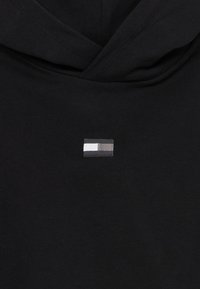 Juodas džemperis su gobtuvu, kuriame krūtinės srityje yra mažas, stačiakampio formos išsiuvinėtas logotipas baltos, šviesiai pilkos ir tamsiai pilkos spalvų. Minkšta, tekstūruota medžiaga.