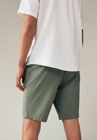 Leichtes, kurzärmeliges weißes Hemd, kombiniert mit grünen Shorts. Die Shorts haben eine glatte Textur und eine simple, lässige Passform.