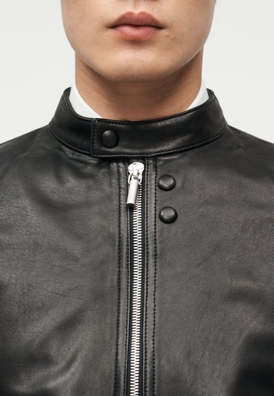 Κοντινό πλάνο ενός ατόμου που φορά μαύρο δερμάτινο μπουφάν με ασημένιο φερμουάρ και κουμπιά τύπου πιέτας στη λαιμόκοψη και την περιοχή του στήθους.