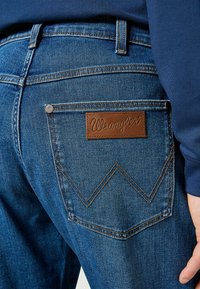 Niebieskie dżinsy mają charakterystyczną skórzaną łatę z logo "Wrangler", kontrastowe szwy oraz wyraźny wzór tylnych kieszeni.