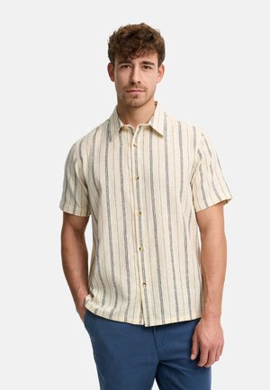 Homme portant une chemise beige à manches courtes à rayures verticales et un pantalon bleu, debout avec une main dans la poche devant un fond uni.