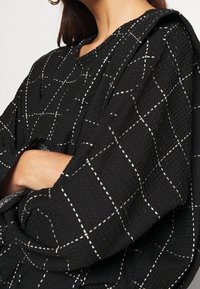 Svart texturerad tygtröja med ett rutmönster i vitt. Har en rundad halsringning och oversized ärmar.