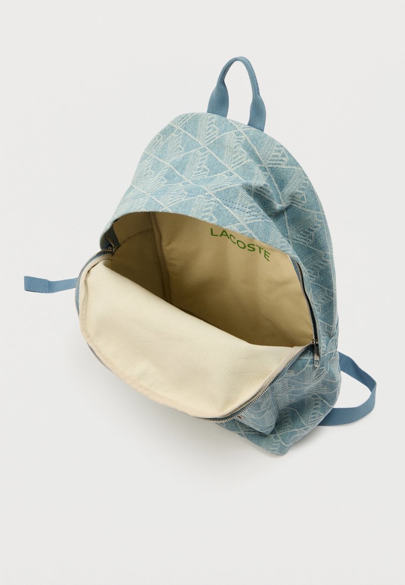 Sac à dos bleu clair à motifs avec compartiment principal ouvert montrant l'intérieur beige et le logo vert Lacoste à l'intérieur.