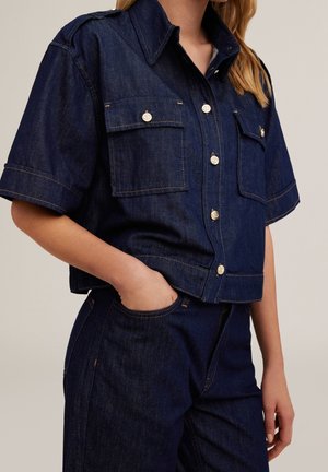Kobieta ubrana w ciemnoniebieską jeansową koszulę z krótkim rękawem zapinaną na guziki z przednimi kieszeniami na klapie oraz dopasowane jeansy z wysokim stanem, ręka w kieszeni.