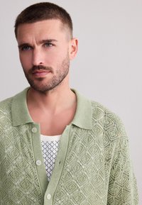 Ljusgrön stickad kofta med ett geometriskt mönster, med krage och knäppning över en vit mönstrad skjorta.