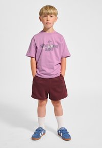 Lavendelfarbenes T-Shirt mit dem Schriftzug "SERVE THE GAME", bordeauxrote Shorts, weiße Knöchelsocken und blaue Klettschuhe mit weißen Akzenten.