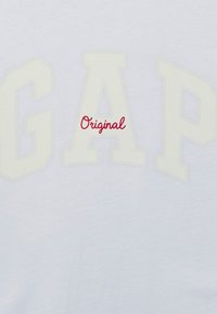 Lyseblå t-shirt med store, prægede hvide bogstaver "GAP" og et rødt kursiv "Original" placeret centralt. Blød stofstruktur.